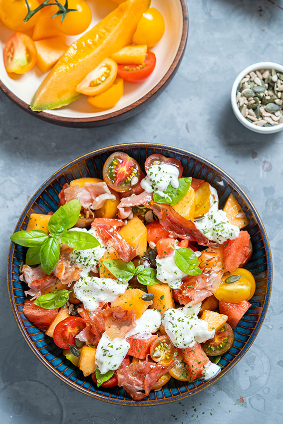 Vue sur un bol contenant de la salade d'été avec fruits, tomates, fromage burrata et jambon. Une assiette posée à côté contient une tranche de melon et des tomates cerises.