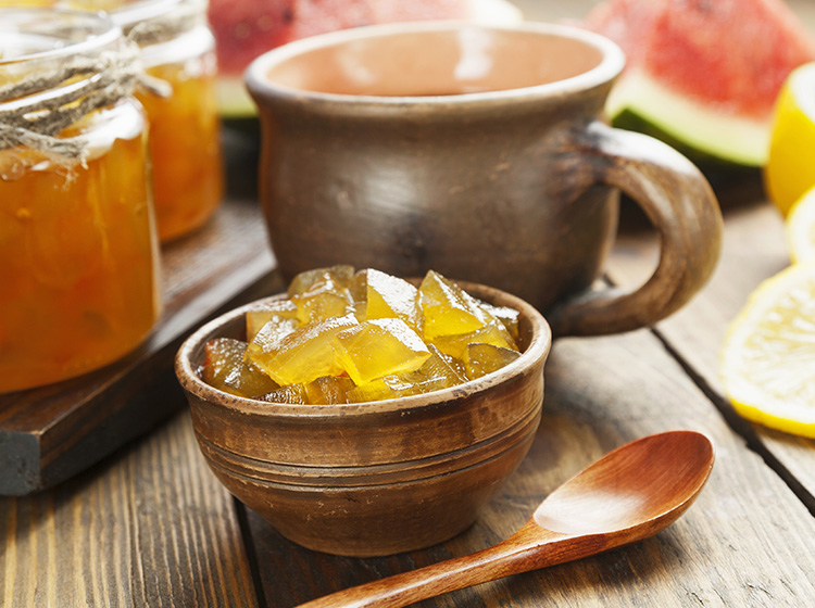 Sur une table est posé un bol rempli de dés confits de pastèque à confire jaune d’or. On distingue en arrière-plan des tranches de pastèque et de citron pour rappeler les ingrédients de la recette.