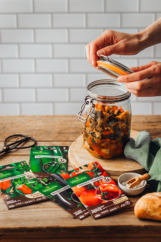 Un homme ferme le bocal de conserve d'aubergines, de poivrons, tomates et courgettes posé sur une table. Des sachets de graines Vilmorin sont posés à côté du bocal.